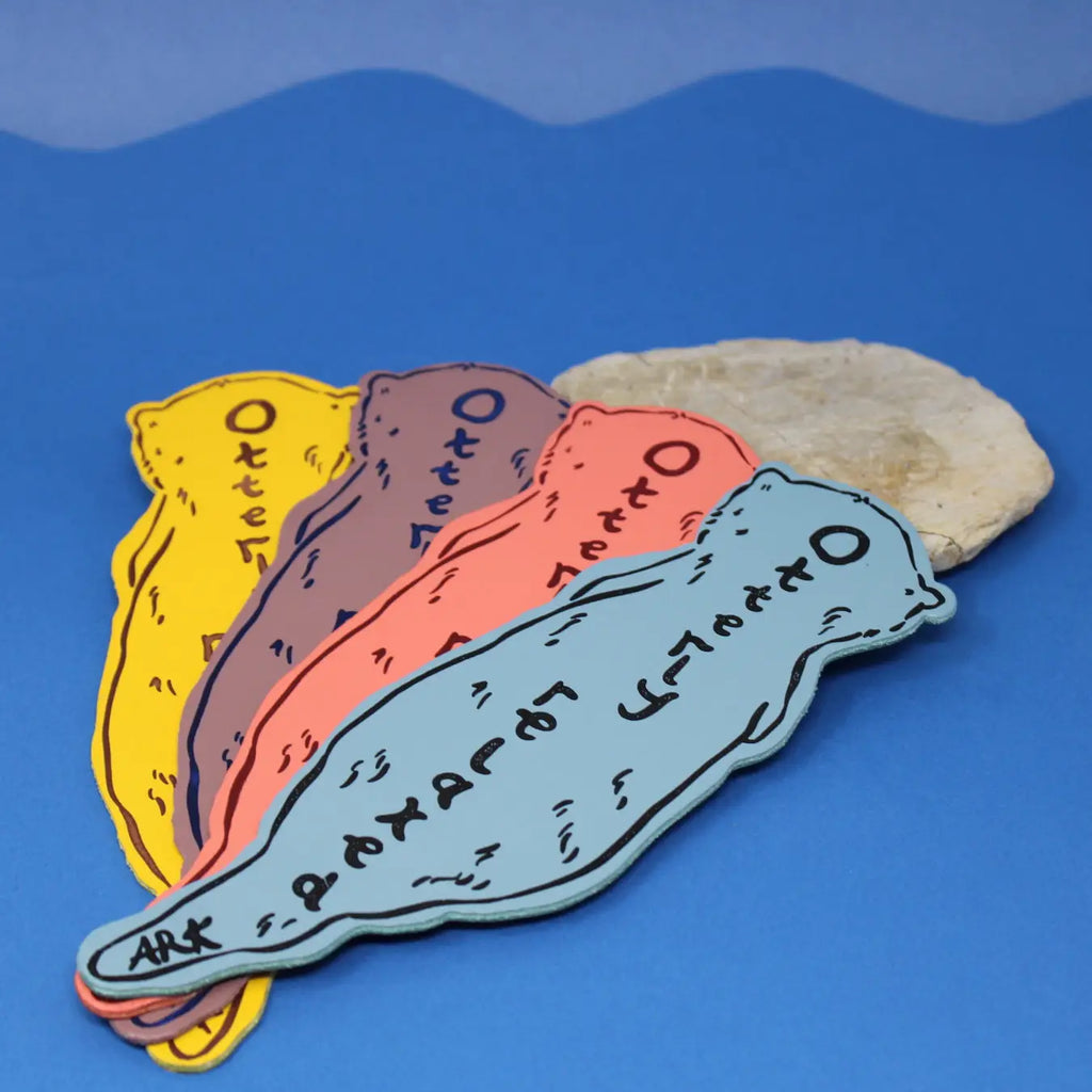 Four otter-shaped bookmarks in blue, coral, dusty lilac, and yellow, each lettered "Ark Otter" along the body, fanned out on a blue background next to a stone.