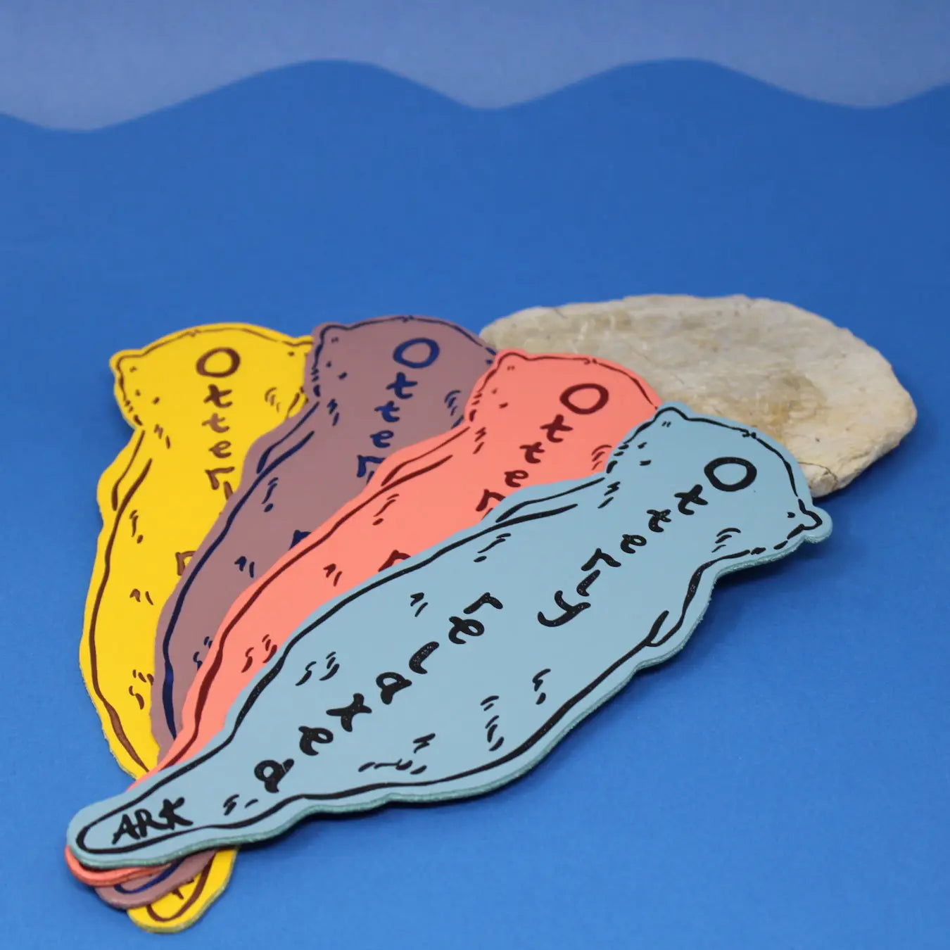 Four otter-shaped bookmarks in blue, coral, dusty lilac, and yellow, each lettered "Ark Otter" along the body, fanned out on a blue background next to a stone.