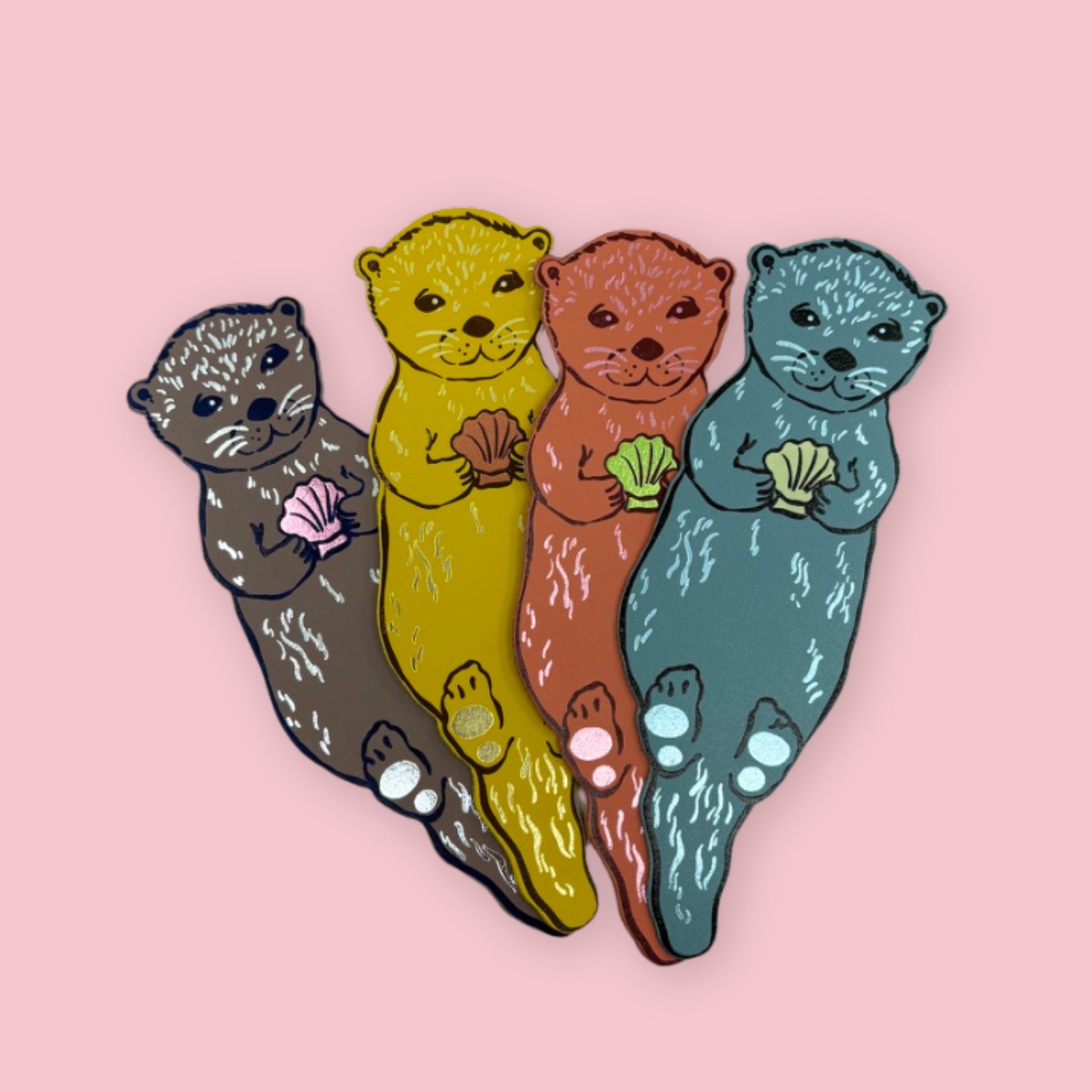 Four leather otter-shaped bookmarks in grey, yellow, orange, and blue on a pale pink background.