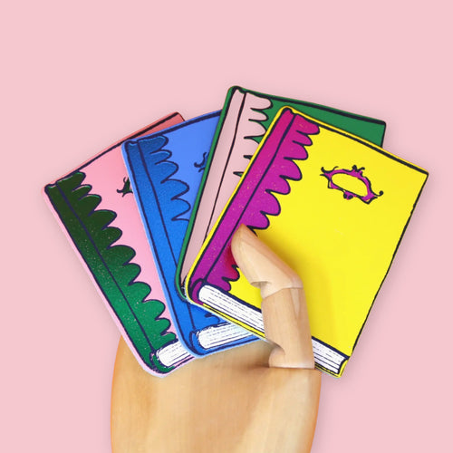 Set of colorful leather coasters by Ark Colour Design illustrated as stacked books, displayed in a wooden mannequin hand against a pastel pink background. The coasters feature bright yellow, blue, pink, and green book covers with whimsical eye motifs and decorative scalloped edges.