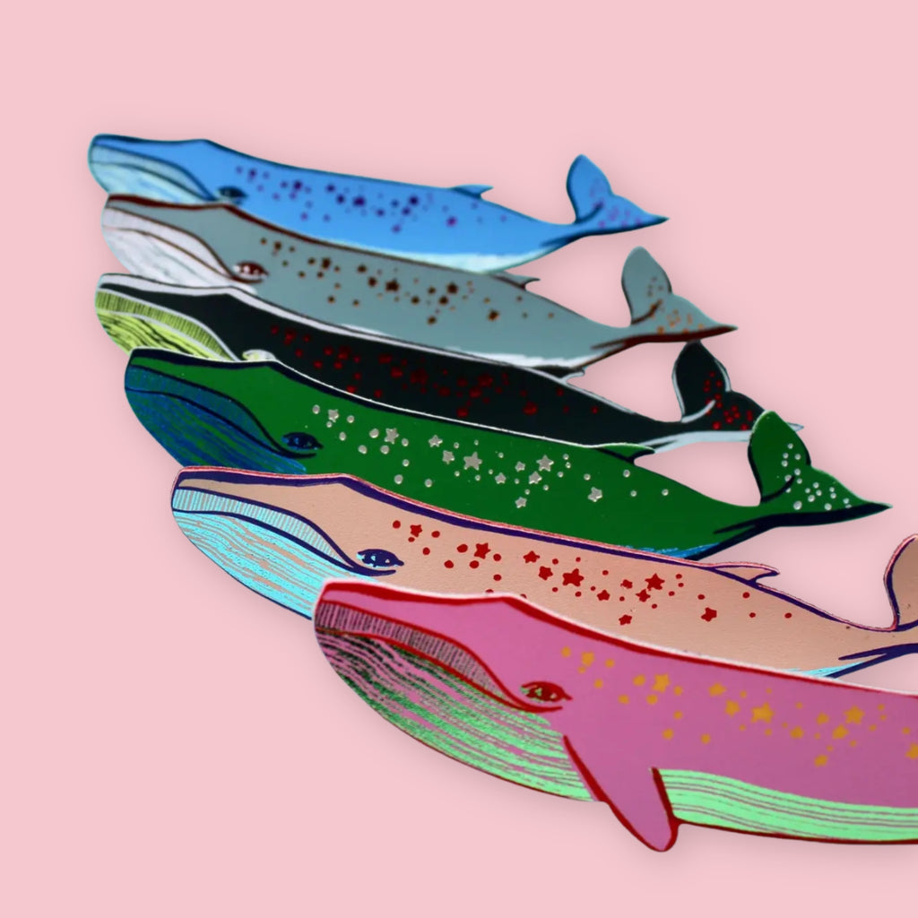 Five whale-shaped bookmarks in blue, grey, green, pale pink, and hot pink, each with stars or dots along the body, fanned out on a pink background.