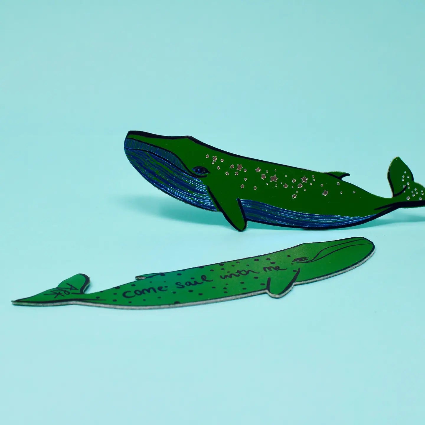 Two green whale-shaped bookmarks, one with star designs along its body and one lettered "Come sail with me," on a light blue background.