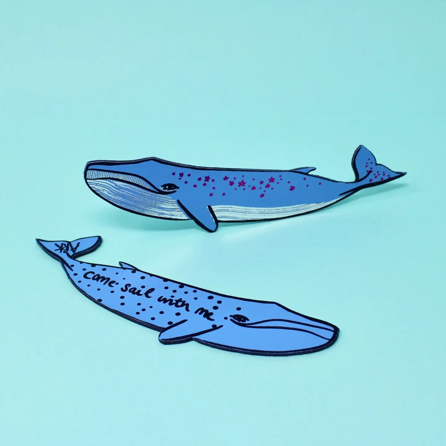Two blue whale-shaped bookmarks, one with star designs along its body and one lettered "Come sail with me," on a light blue background.