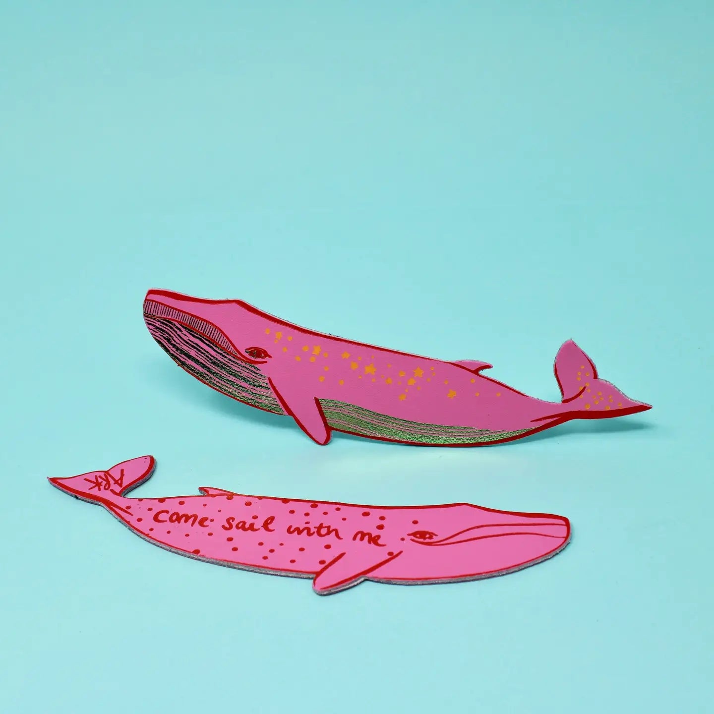 Two hot pink whale-shaped bookmarks, one with star designs along its body and one lettered "Come sail with me," on a light blue background.