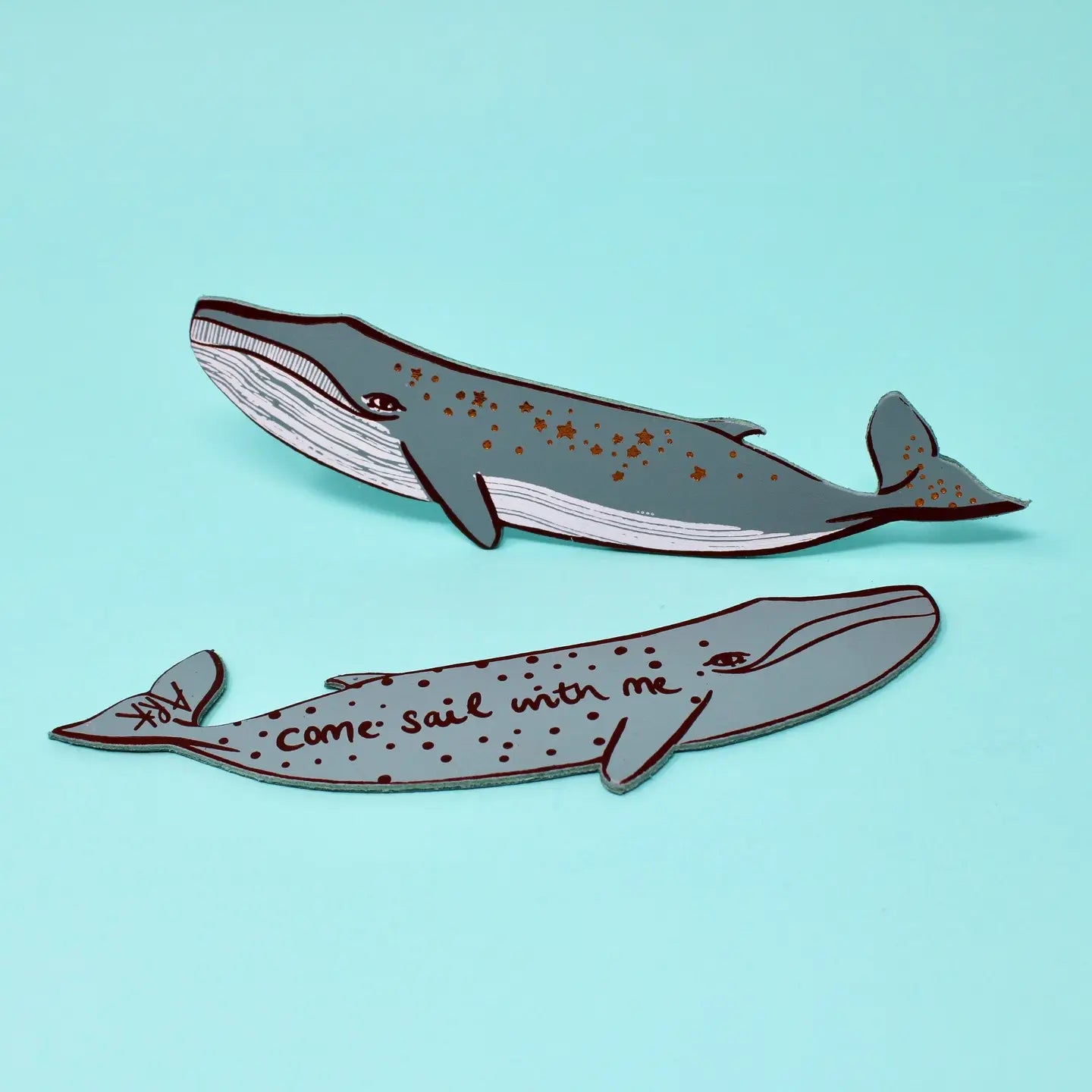 Two grey whale-shaped bookmarks, one with star designs along its body and one lettered "Come sail with me," on a light blue background.