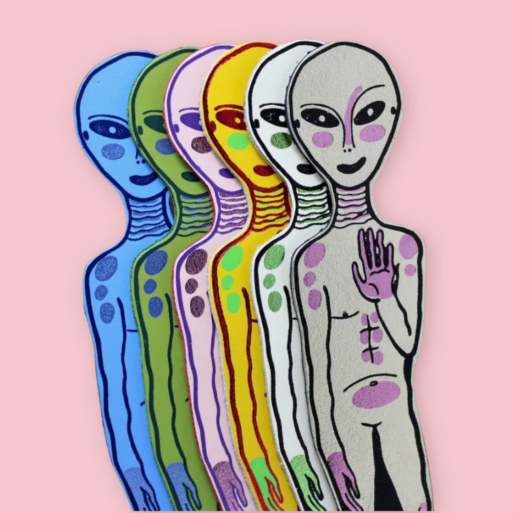 Six alien-shaped bookmarks in blue, green, pink, yellow, white, and gold, each with raised hand and spotted body, fanned out on a pink background
