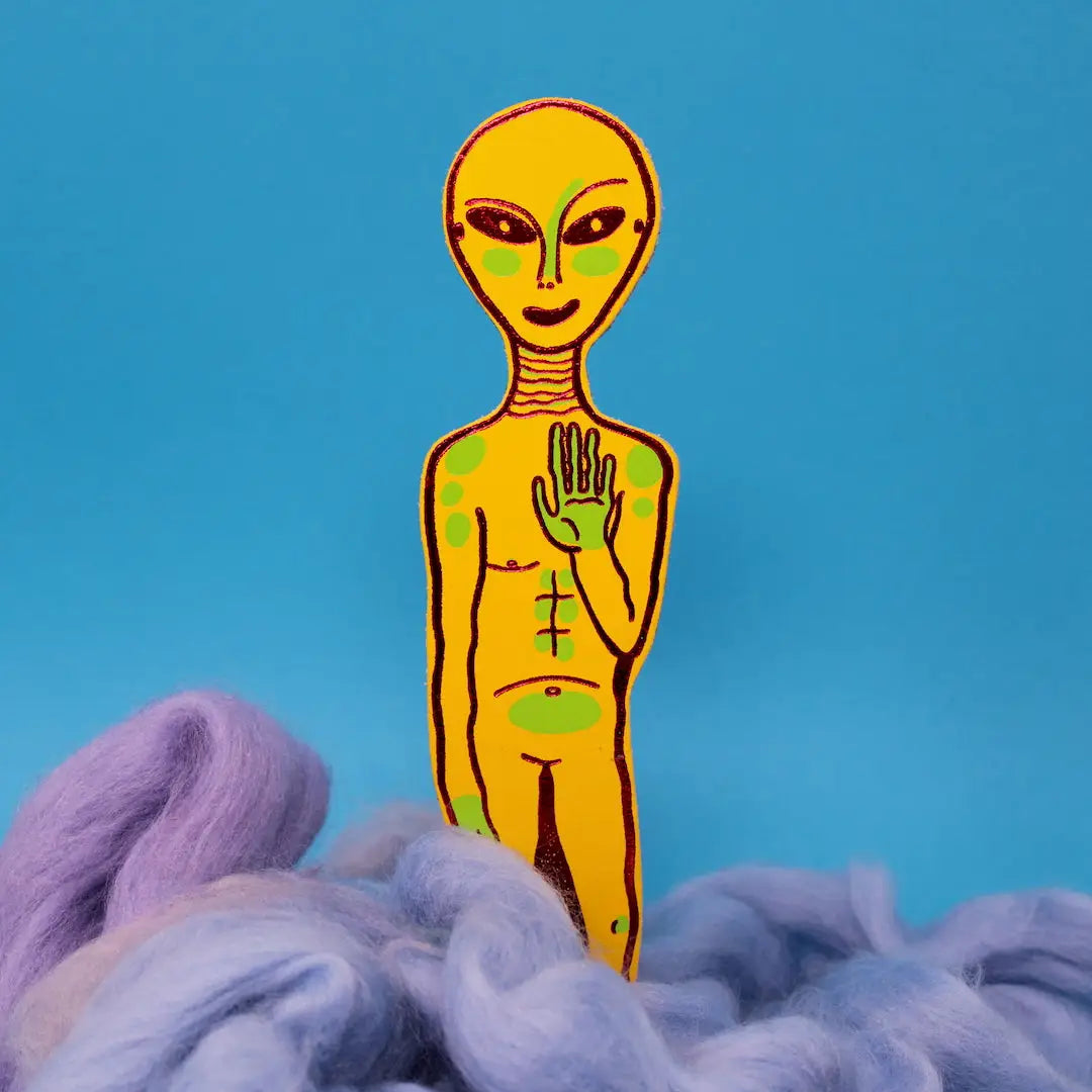 Yellow alien-shaped bookmark with raised hand and orange feet, standing in blue and lavender wool, on a yellow background.