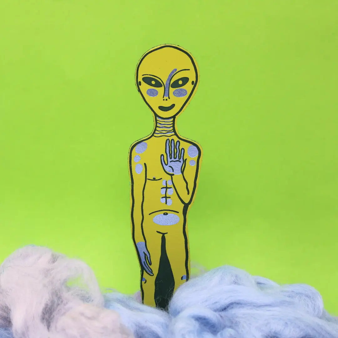 Apple green alien-shaped bookmark with raised hand and orange feet, standing in blue and lavender wool, on a yellow background.