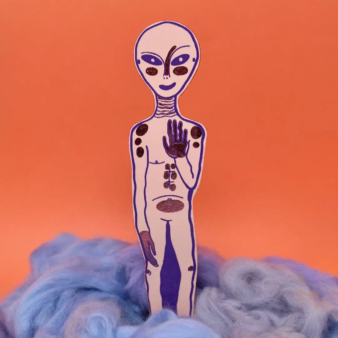Pink alien-shaped bookmark with raised hand and orange feet, standing in blue and lavender wool, on a yellow background.
