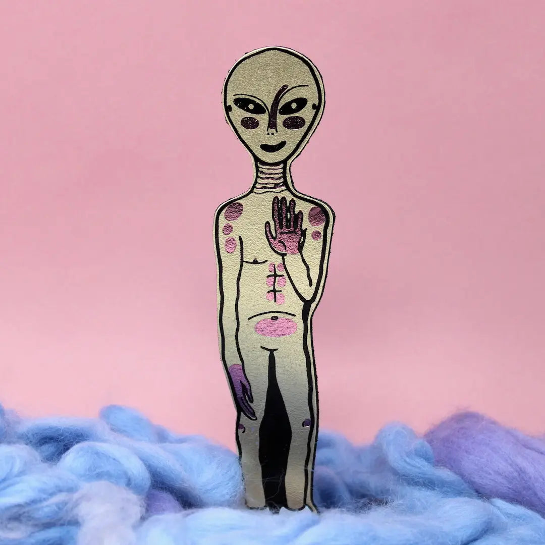 Gold alien-shaped bookmark with raised hand and orange feet, standing in blue and lavender wool, on a yellow background.