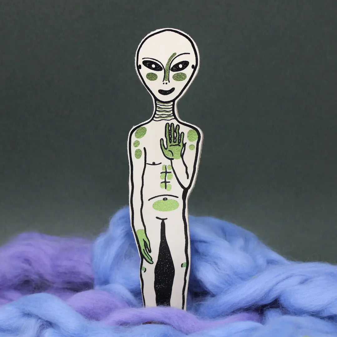 Cream alien-shaped bookmark with raised hand and orange feet, standing in blue and lavender wool, on a yellow background.
