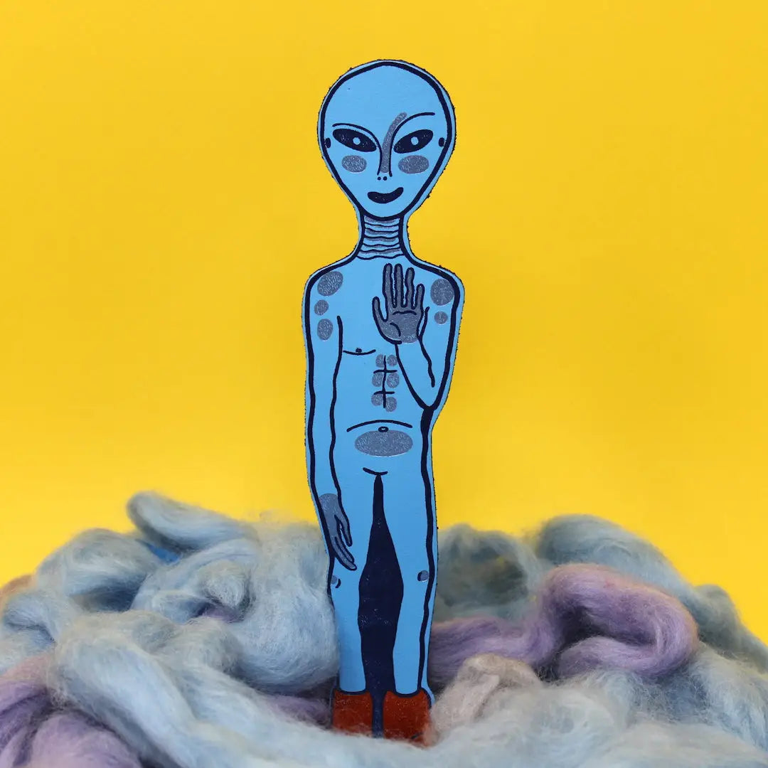 Blue alien-shaped bookmark with raised hand and orange feet, standing in blue and lavender wool, on a yellow background.