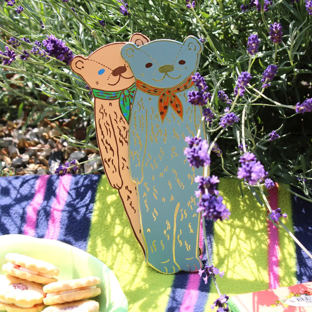 Two Ark Colour Design leather bookmarks shaped like bears, shown standing among lavender flowers on a picnic blanket.