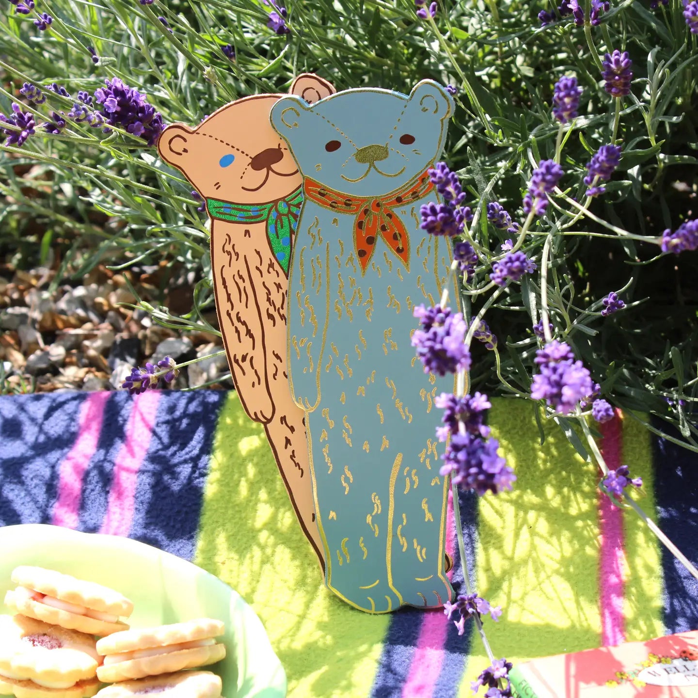 Two Ark Colour Design leather bookmarks shaped like bears, shown standing among lavender flowers on a picnic blanket.