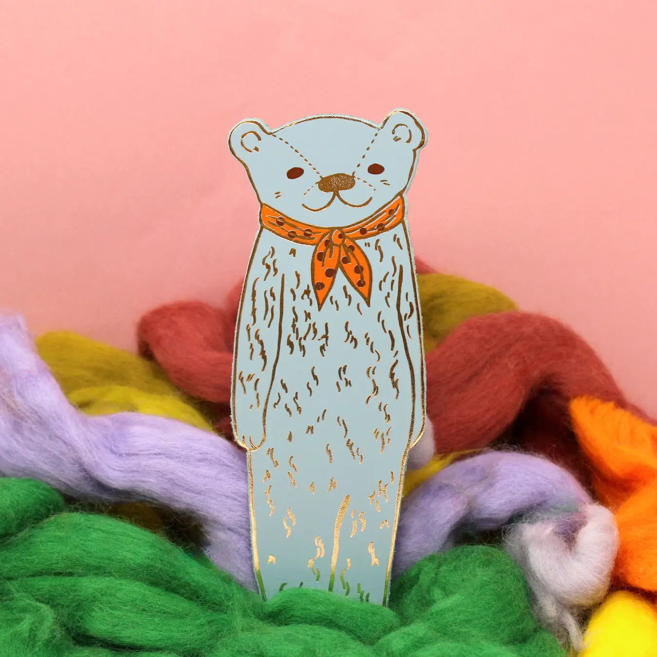 Ark Colour Design leather bookmark in blue shaped like a bear wearing a red scarf, shown standing among colorful wool on a pink background.