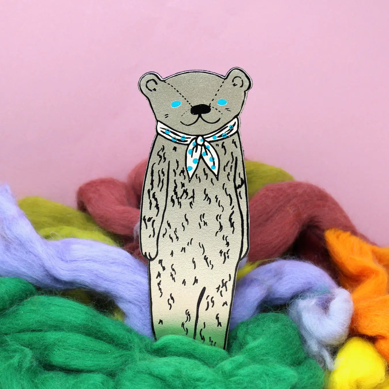 Ark Colour Design leather bookmark in gold shaped like a bear wearing a red scarf, shown standing among colorful wool on a pink background