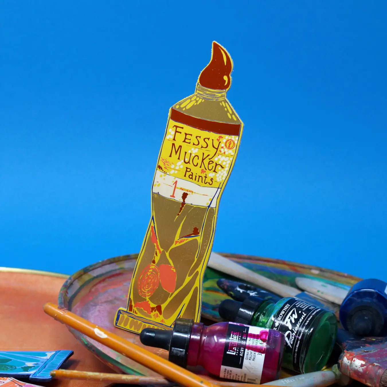 Yellow Ark Colour Design paint tube bookmark with “Fessy Mucker Paints” lettering, shown with paintbrushes and art supplies on a blue background