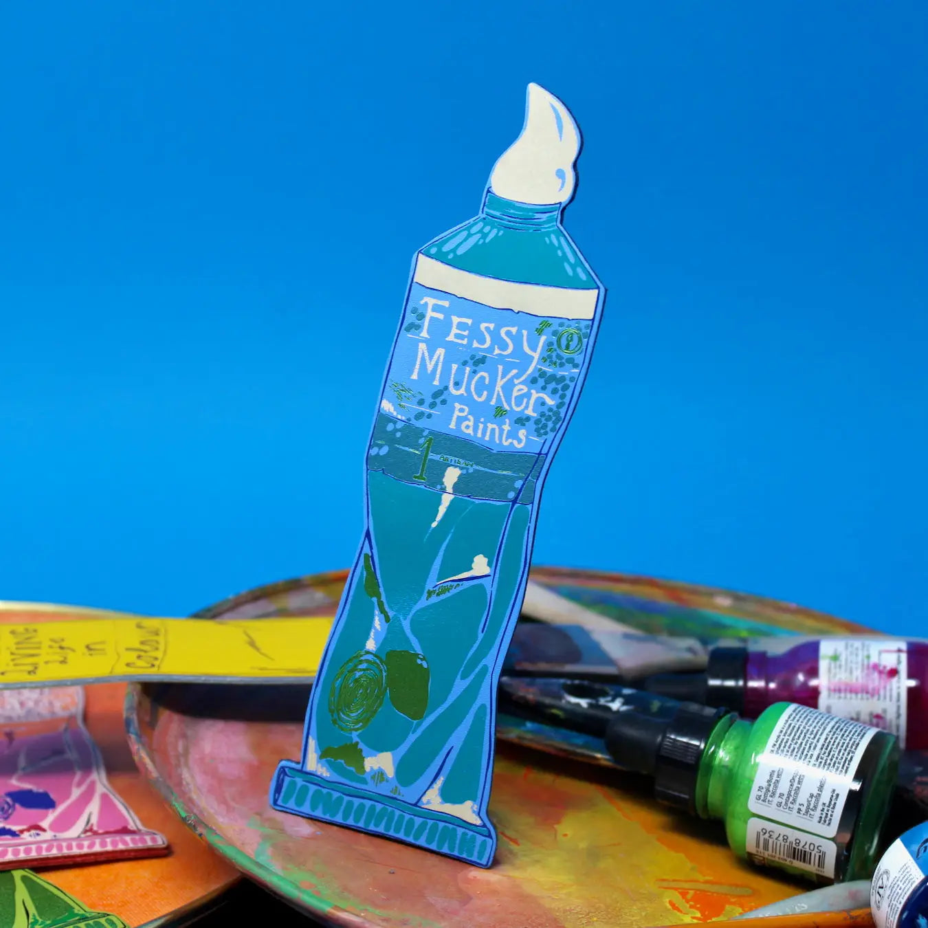 Blue Ark Colour Design paint tube bookmark with “Fessy Mucker Paints” lettering, shown with paintbrushes and art supplies on a blue background