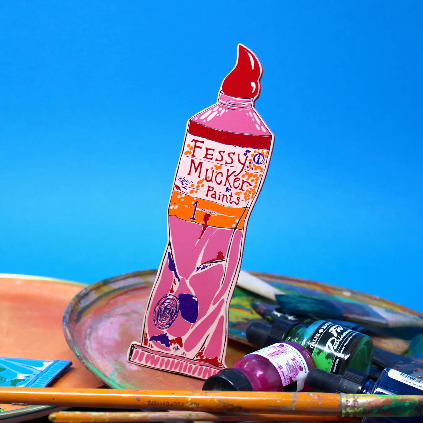 Pink Ark Colour Design paint tube bookmark with “Fessy Mucker Paints” lettering, shown with paintbrushes and art supplies on a blue background