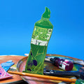 Green Ark Colour Design paint tube bookmark with “Fessy Mucker Paints” lettering, shown with paintbrushes and art supplies on a blue background