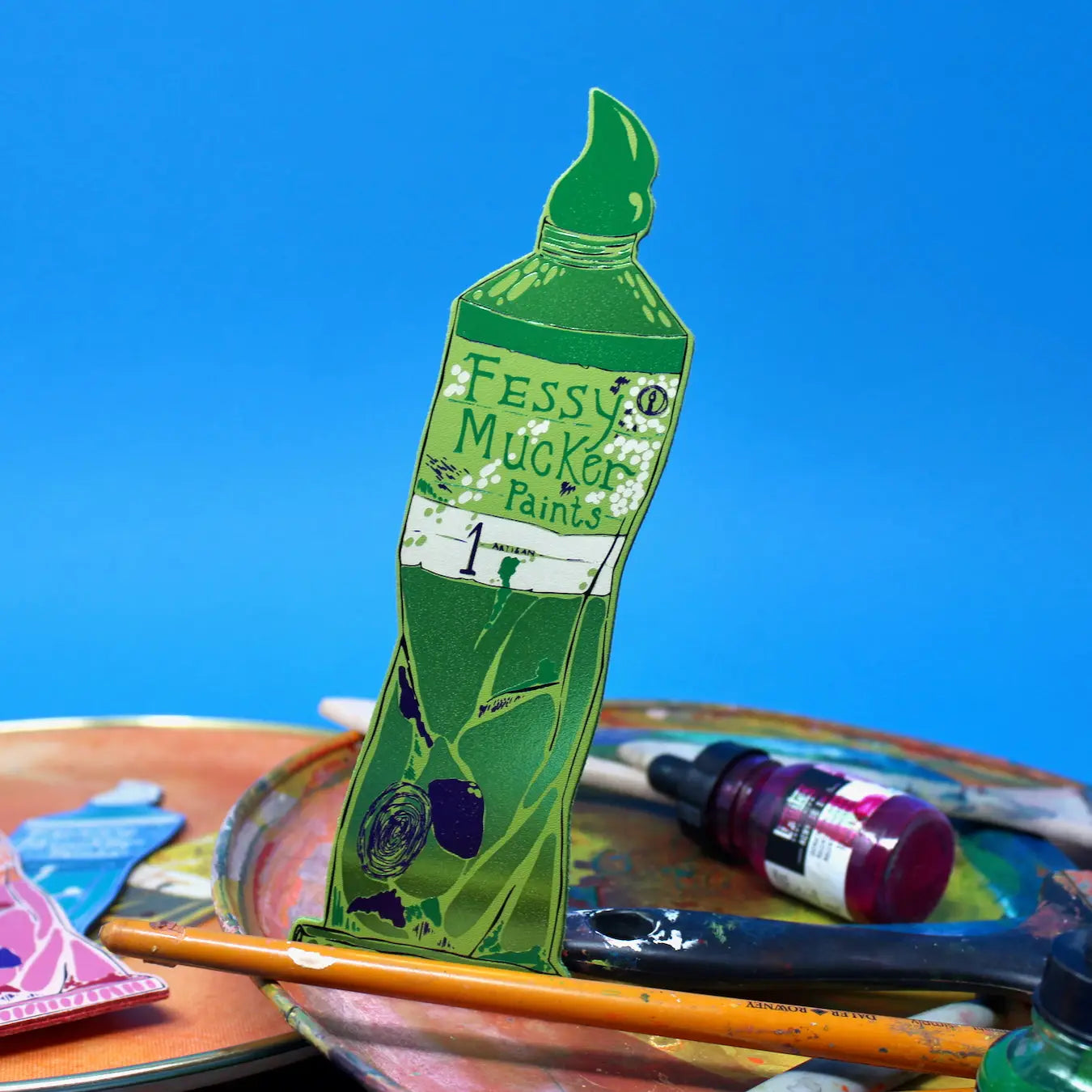 Green Ark Colour Design paint tube bookmark with “Fessy Mucker Paints” lettering, shown with paintbrushes and art supplies on a blue background