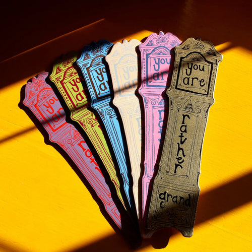 Set of colorful Ark Colour Design leather bookmarks shaped like grandfather clocks with the text 'You are rather grand', fanned out in sunlight on a yellow background.