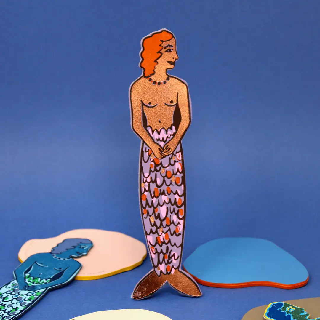 Ark Colour Design leather bookmark shaped like a mermaid, shown standing on a blue background with other mermaid bookmarks nearby.