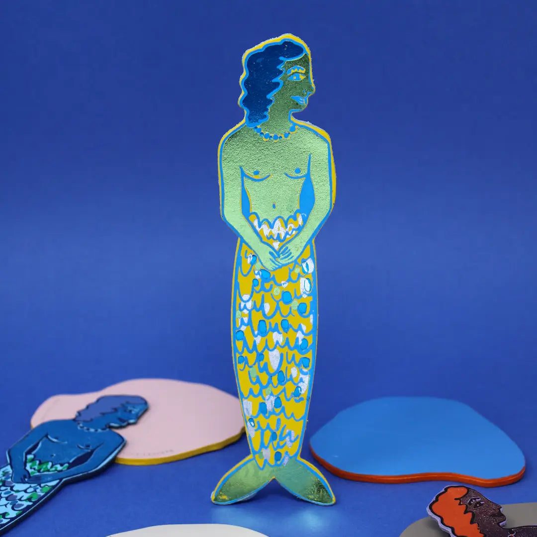 Ark Colour Design leather bookmark shaped like a mermaid, shown standing on a blue background with other mermaid bookmarks nearby.