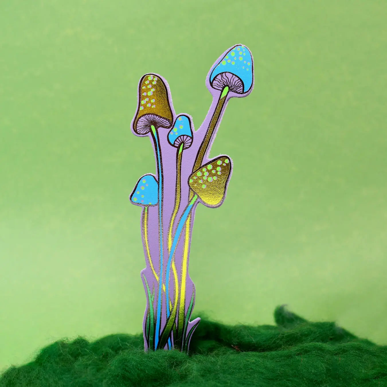 Blue and lilac spotted mushroom-shaped bookmark rising from yellow moss, on a green background.