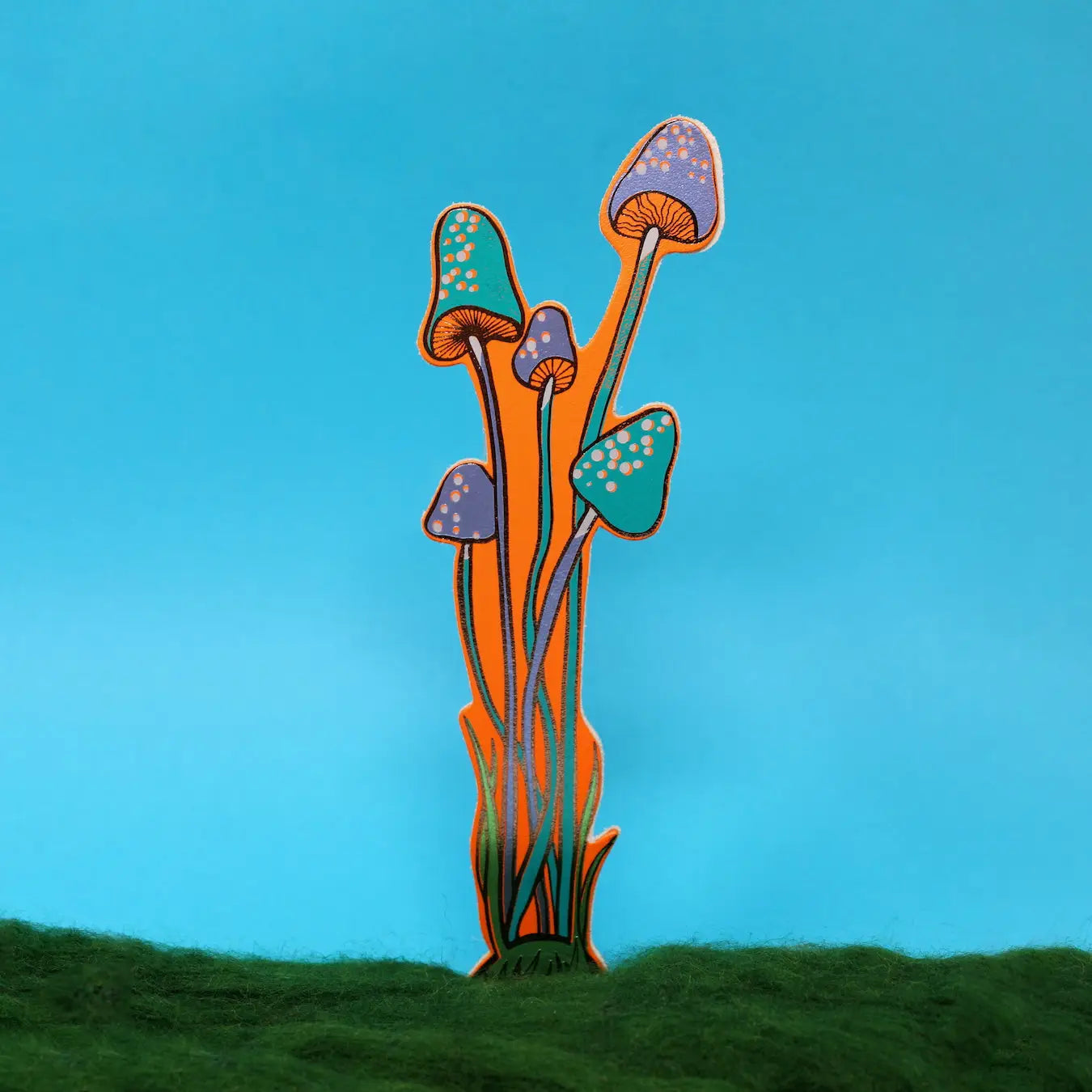 Blue and orange spotted mushroom-shaped bookmark rising from yellow moss, on a green background.