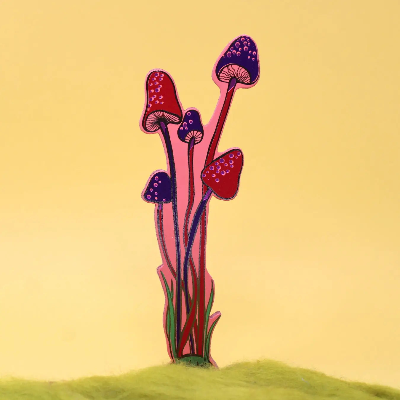 Blue and pink spotted mushroom-shaped bookmark rising from yellow moss, on a green background.