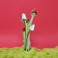Green, red and white spotted mushroom-shaped bookmark rising from yellow moss, on a green background.