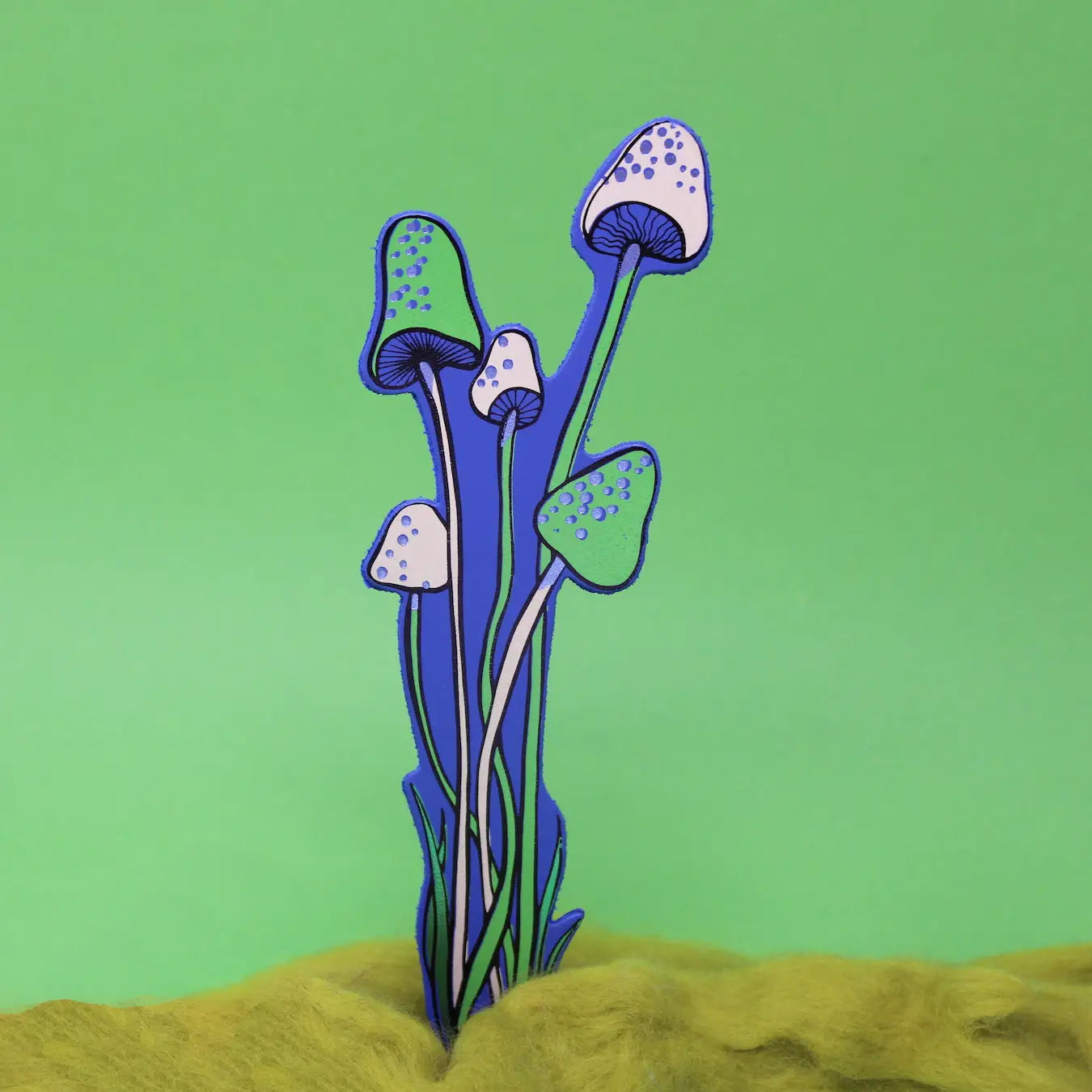 Blue and white spotted mushroom-shaped bookmark rising from yellow moss, on a green background.