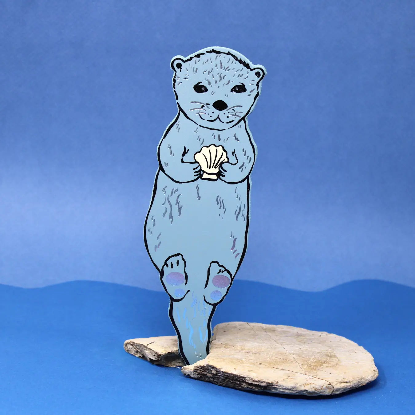Light blue otter-shaped bookmark holding a clam shell, standing on a piece of driftwood against a blue background.