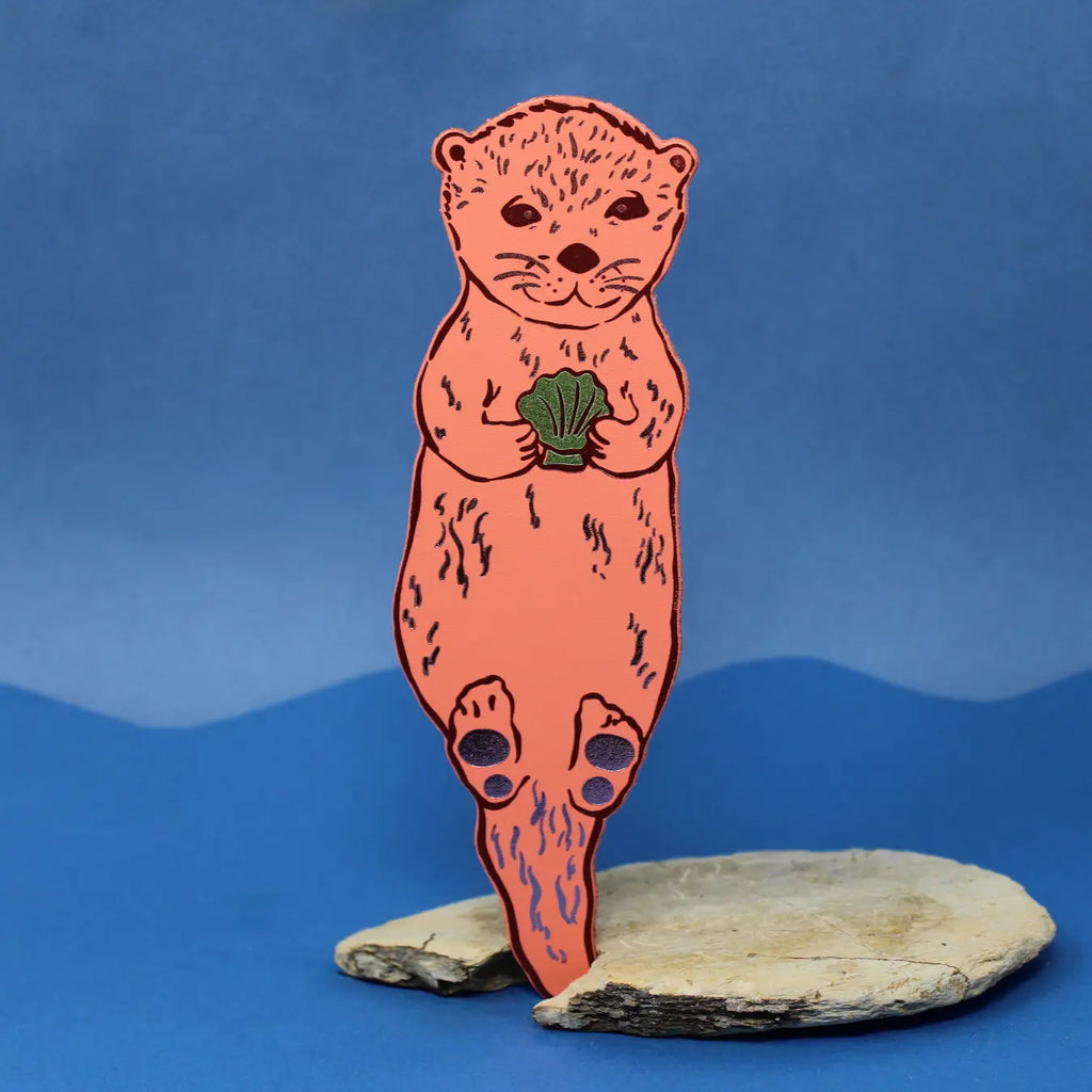 Apricot otter-shaped bookmark holding a clam shell, standing on a piece of driftwood against a blue background.