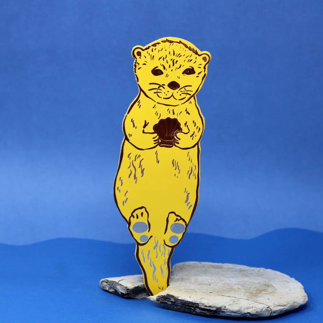 Yellow otter-shaped bookmark holding a clam shell, standing on a piece of driftwood against a blue background.