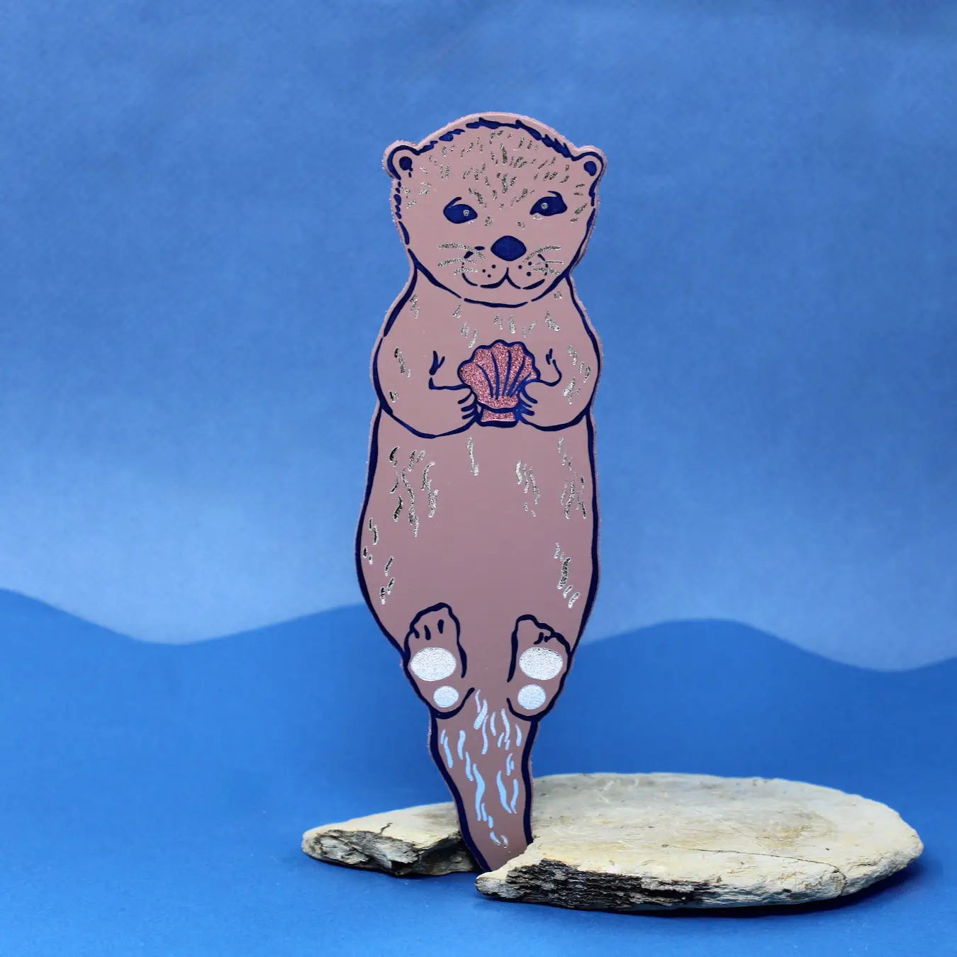Dusty lilac otter-shaped bookmark holding a clam shell, standing on a piece of driftwood against a blue background.