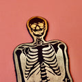 Ark Colour Design skeleton bookmark in black and gold, shown close up on an orange background.