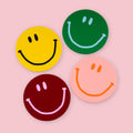Four round leather smiley face coasters by Ark Colour Design in yellow, green, burgundy, and pink on a pastel pink background.