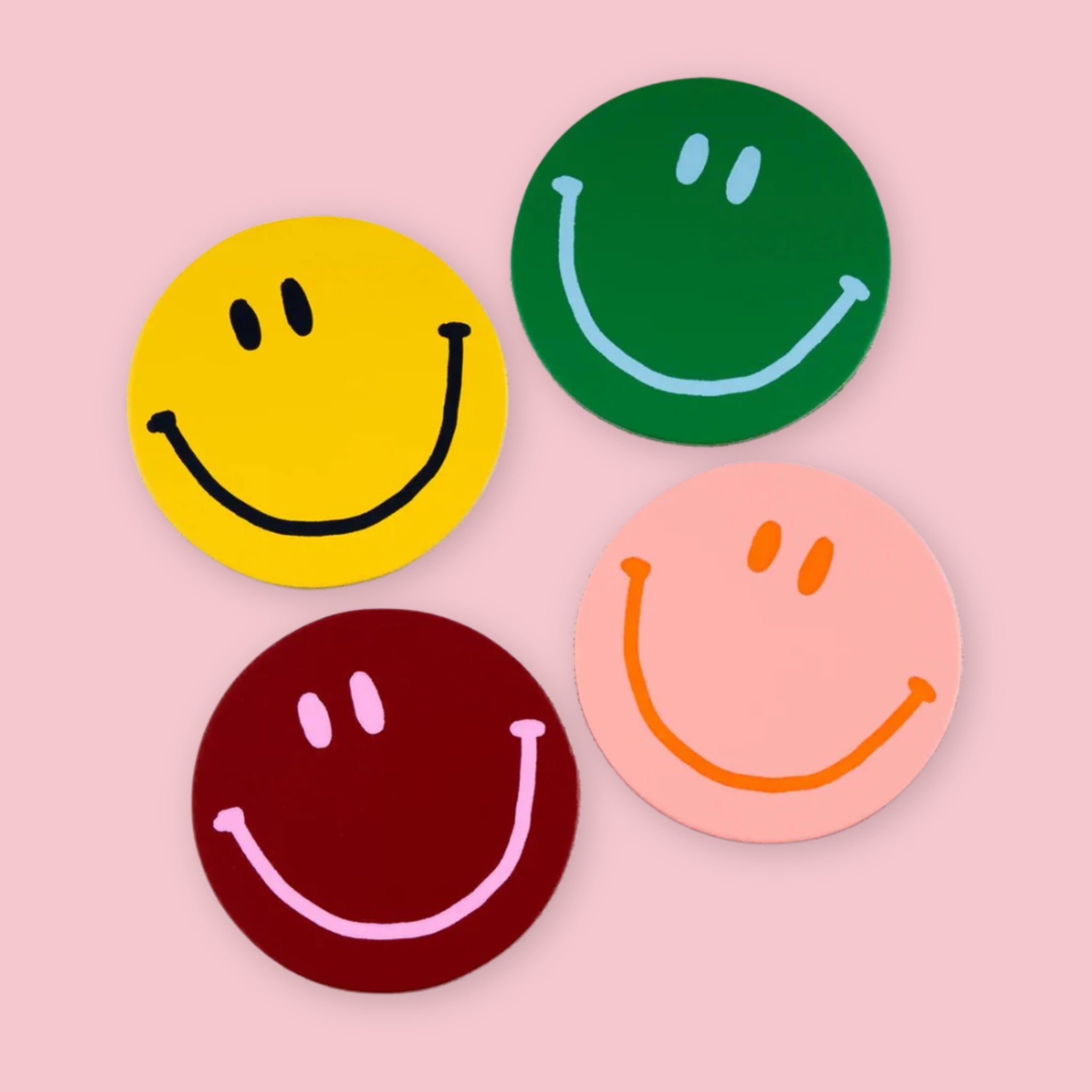 Four round leather smiley face coasters by Ark Colour Design in yellow, green, burgundy, and pink on a pastel pink background.