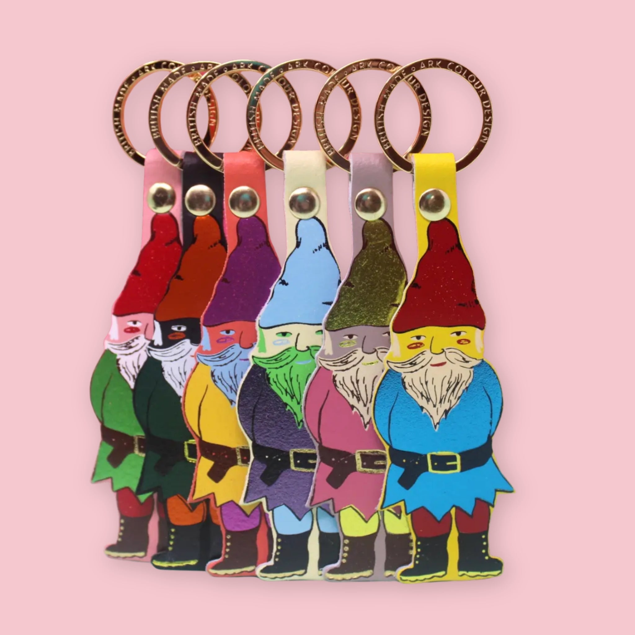 Ark Colour Design gnome keychains in six colorful styles, arranged in a row with gold key rings on a pink background.