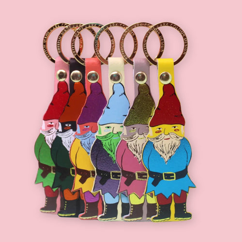 Ark Colour Design gnome keychains in six colorful styles, arranged in a row with gold key rings on a pink background.