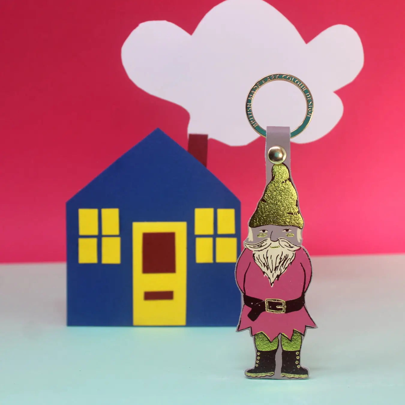 Ark Colour Design gnome keychain in nude with gold ring, shown in front of a colorful house and cloud backdrop.