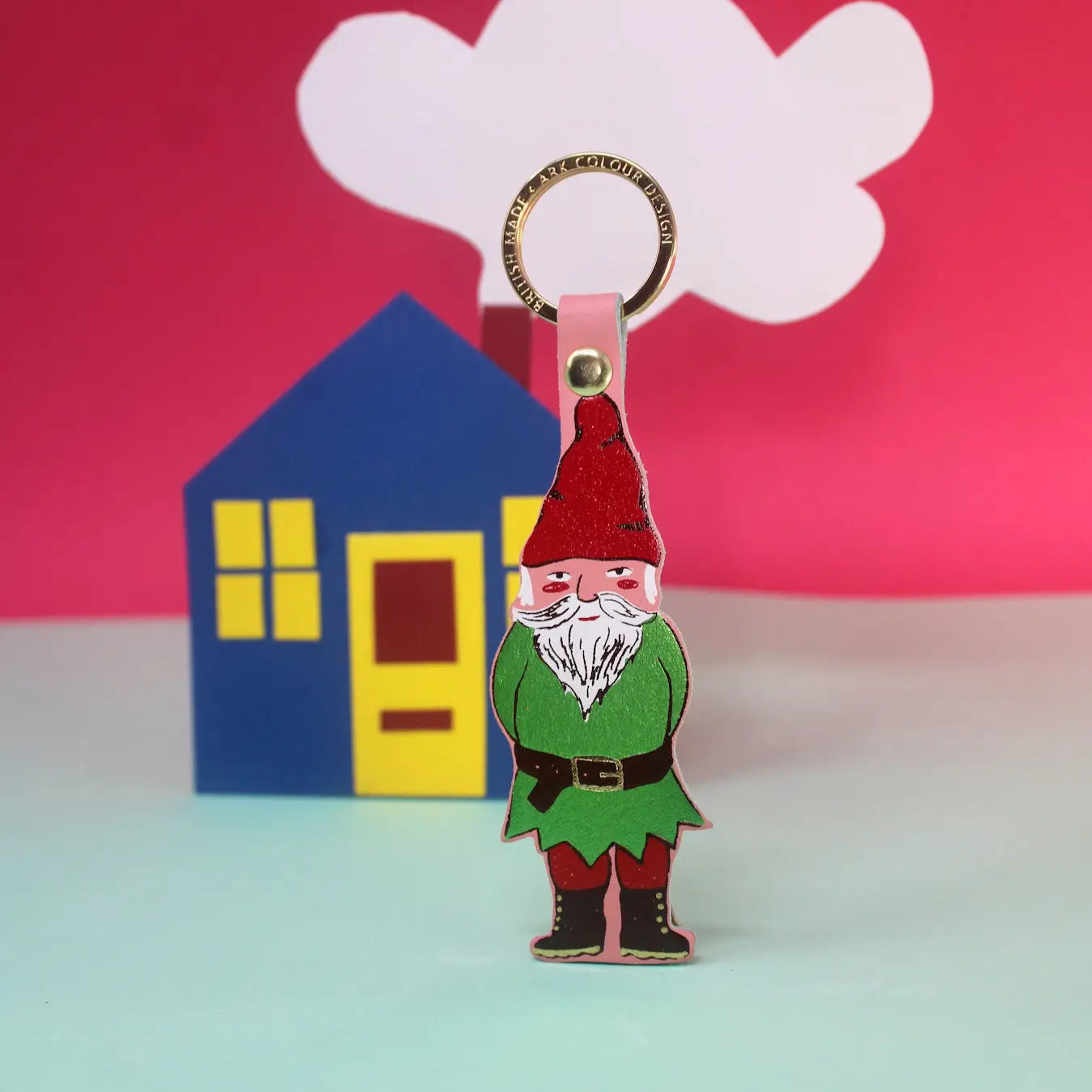 Ark Colour Design gnome keychain in pale pink with gold ring, shown in front of a colorful house and cloud backdrop.