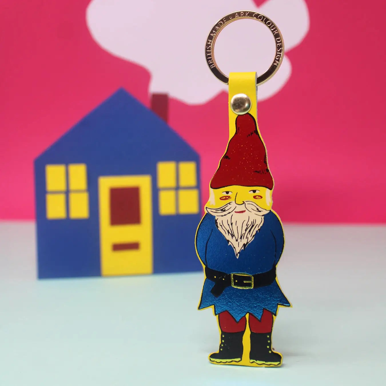 Ark Colour Design gnome keychain in yellow with gold ring, shown in front of a colorful house and cloud backdrop.
