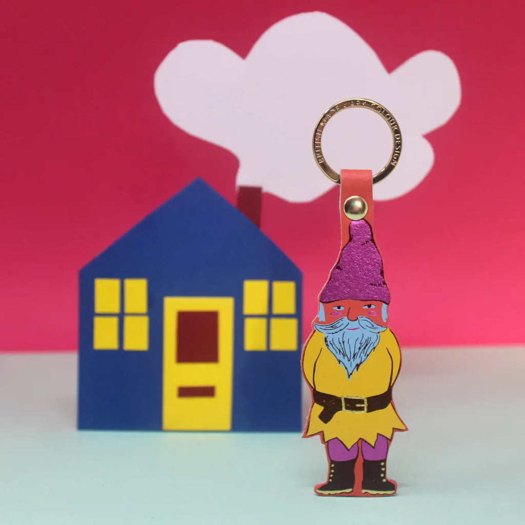 Ark Colour Design gnome keychain in coral with gold ring, shown in front of a colorful house and cloud backdrop.
