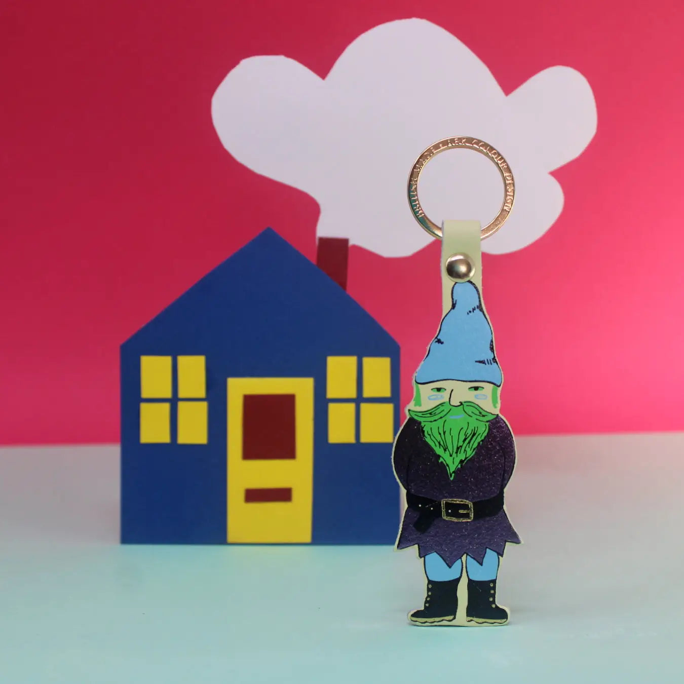 Ark Colour Design gnome keychain in cream with gold ring, shown in front of a colorful house and cloud backdrop.