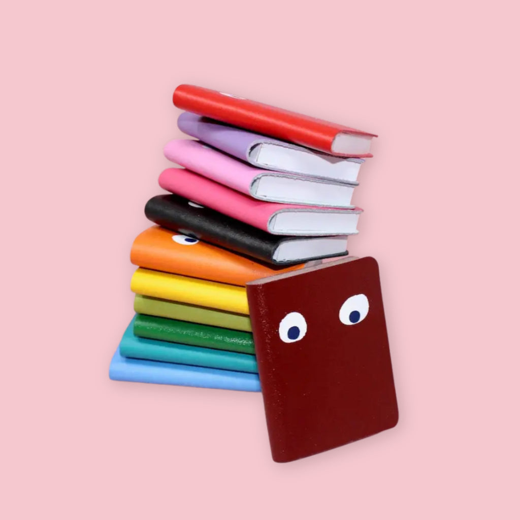 Stack of Ark Colour Design leather googly eye notebooks in multiple colors including red, lilac, pink, black, orange, yellow, green, and turquoise on a pink background