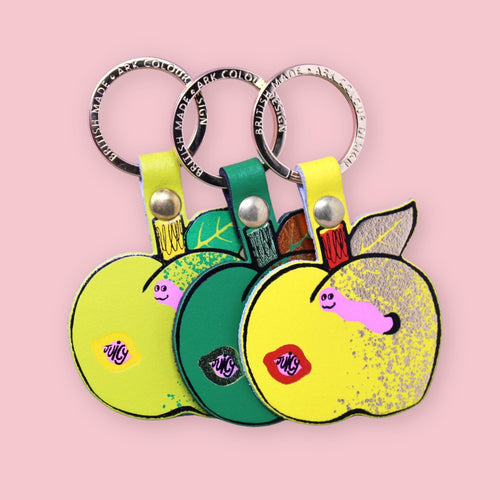 Ark Colour Design apple keychains in yellow and green colorways, shown with gold rings on a pink background.