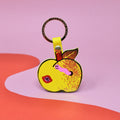 Ark Colour Design apple keychain with yellow strap, gold ring, and illustrated red apple design on a pink and red background.